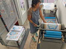 Load image into Gallery viewer, An Asian woman artist operating an old press, printing the sash block in the "I Deserve Love and Comfort" print. A large printing rack is behind her with some finished prints.
